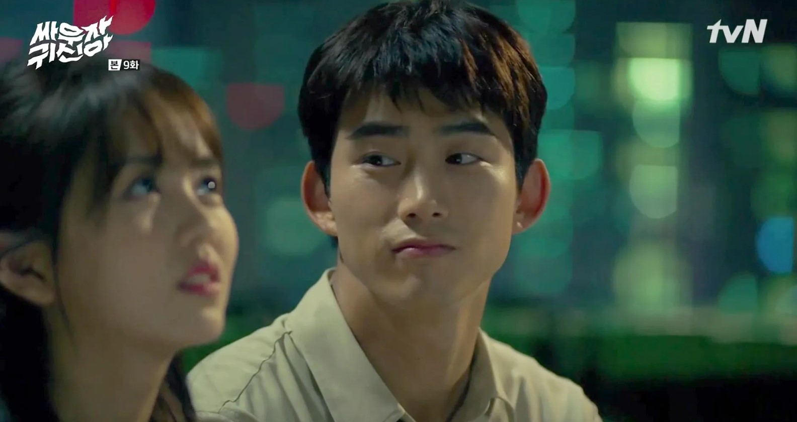 A scene from the Korean drama Let’s Fight the Ghost, showing the two main characters sitting next to each other. The female character looks upwards, while the male character glances at her with a playful or curious expression. The background features a soft, blurred cityscape with green and blue lights.