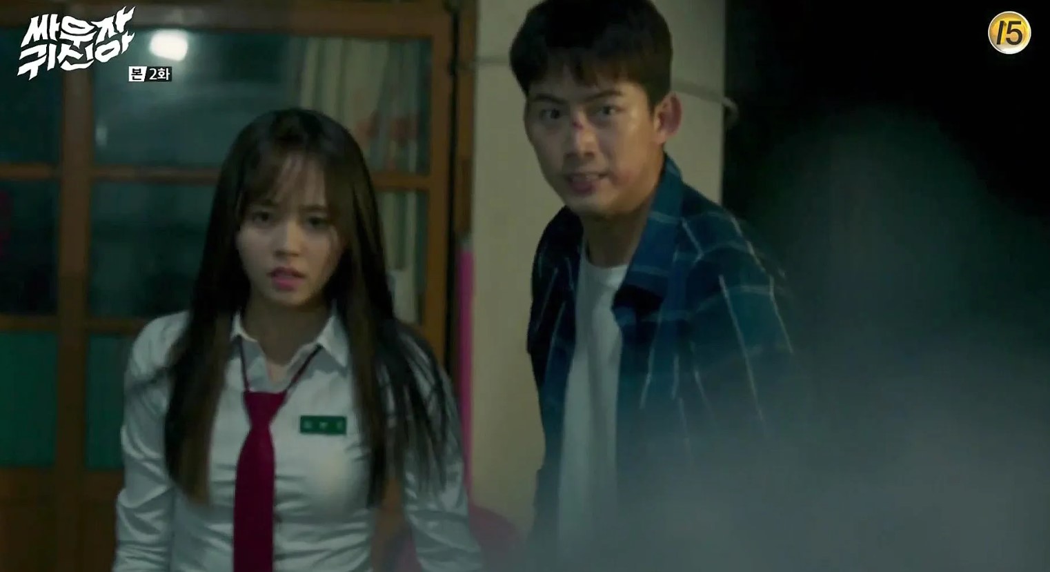 A tense scene from the Korean drama Let’s Fight the Ghost, where the two main characters, a male and female character, are looking alarmed. The female character is dressed in a school uniform, and the male character, with a visible injury on his face, is wearing a checkered shirt. They appear to be in a dimly lit room, possibly preparing to face a ghostly threat. 