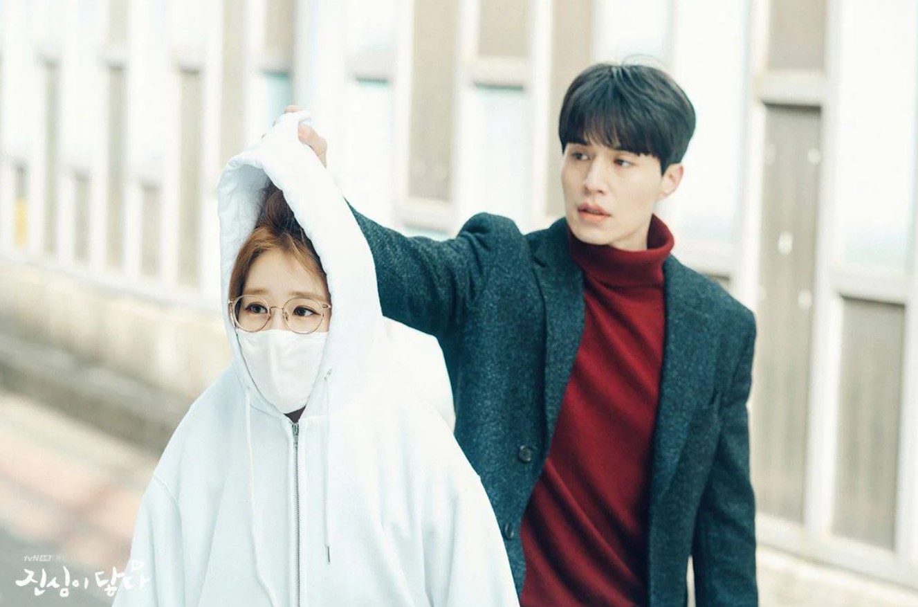 Main characters from the drama Touch Your Heart – the man in a red turtleneck and grey coat lifting the woman's hoodie as she wears glasses and a face mask, both standing outside. 