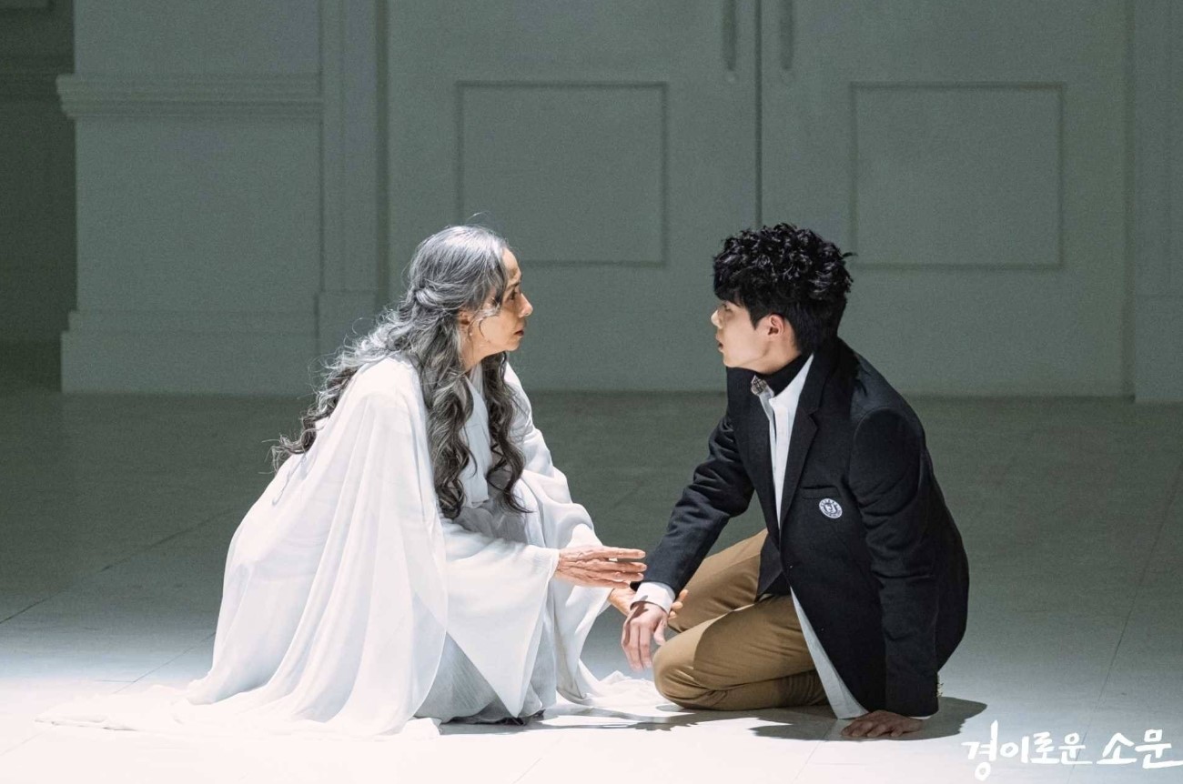 Scene from Uncanny Counter Season 1 – a dramatic moment with a woman in white and a young boy in a suit, both sitting on the floor, facing each other with a deep, emotional exchange.
