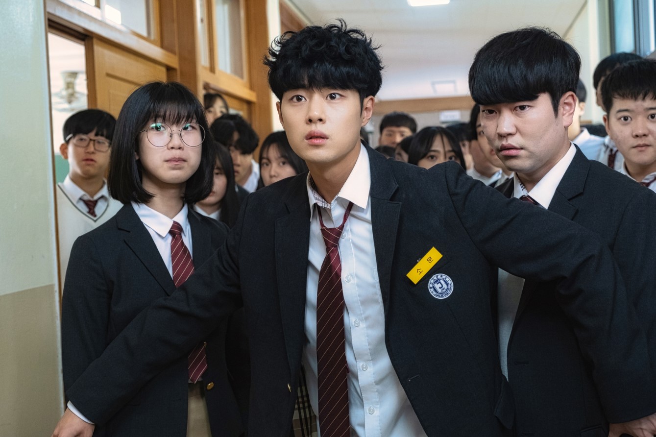 Main character from Uncanny Counter Season 1 – a young man in a school uniform, looking concerned and leading a group of classmates through a hallway.