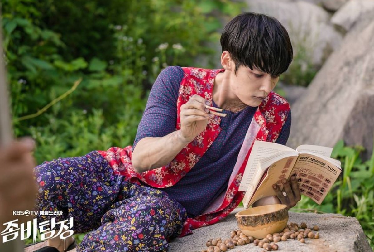 Zombie Detective K-Drama scene with the male lead reading a book while eating, dressed in colorful attire in a natural outdoor setting.