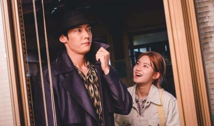 Zombie Detective K-Drama scene with the male and female leads sharing a light-hearted moment, as the male lead adjusts his coat while the female lead smiles.