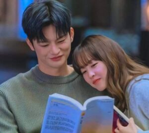 Doom at Your Service scene with the male and female leads smiling together while reading a book in a cozy indoor setting