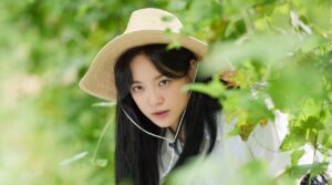 The female lead from "Drinking Solo" in a serene outdoor setting, wearing a straw hat and peeking through greenery, capturing a moment of curiosity and charm.
