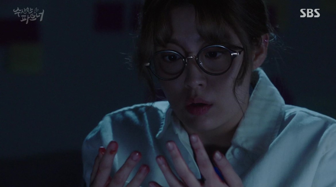 A tense scene from Suspicious Partner, showing the female lead in a moment of shock or fear, staring at her hands with wide eyes, as she processes a critical or alarming situation in a dimly lit room.