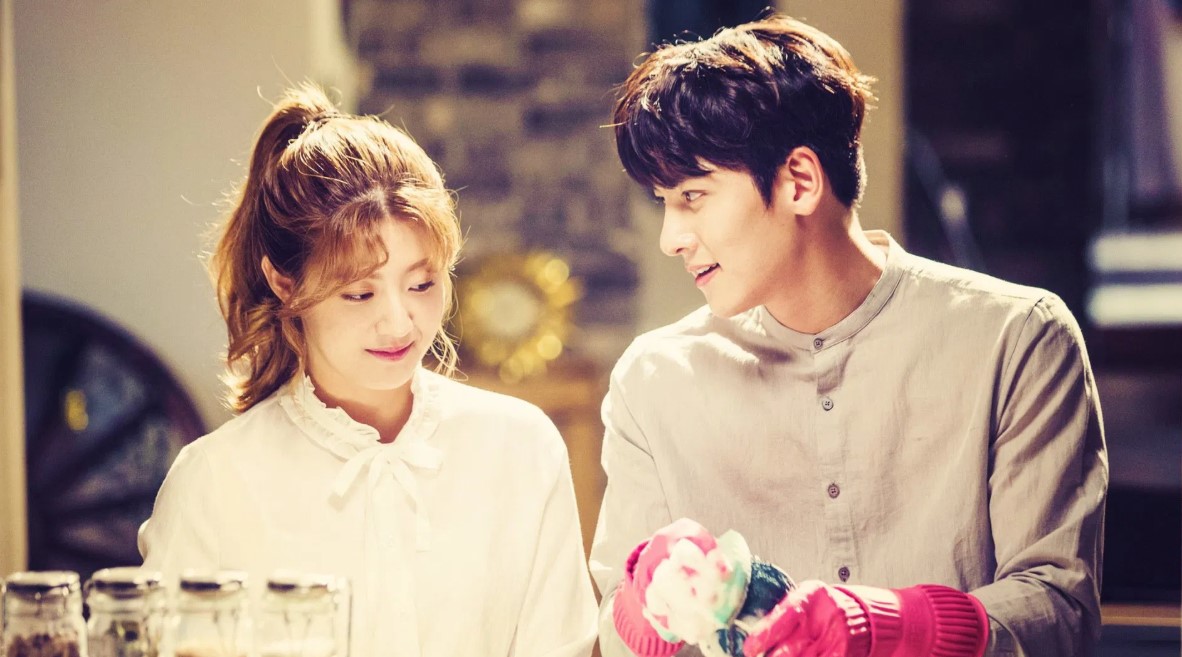 A joyful couple smiling at each other while working together in a cozy kitchen, with the man wearing pink rubber gloves and holding a colorful dish, creating a warm and playful atmosphere. 