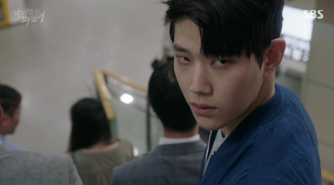 A dramatic moment from Suspicious Partner, featuring the male lead looking over his shoulder with a serious and intense expression, as he stands on an escalator surrounded by other people. 