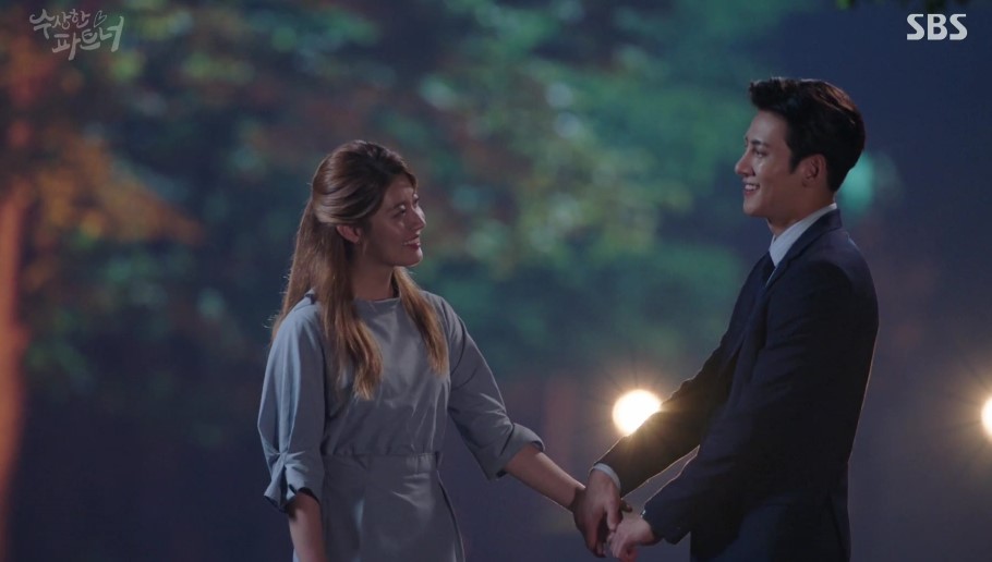 A romantic scene from Suspicious Partner, featuring the male and female leads holding hands and smiling at each other, set against a beautifully lit outdoor background, capturing a joyful and tender moment.
