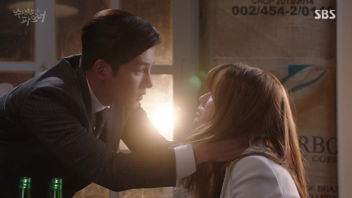 .A tense moment from Suspicious Partner, where the male lead, dressed in a suit, is holding the female lead's face in a dramatic gesture, with the warm light of the setting sun highlighting the intensity of the scene. 