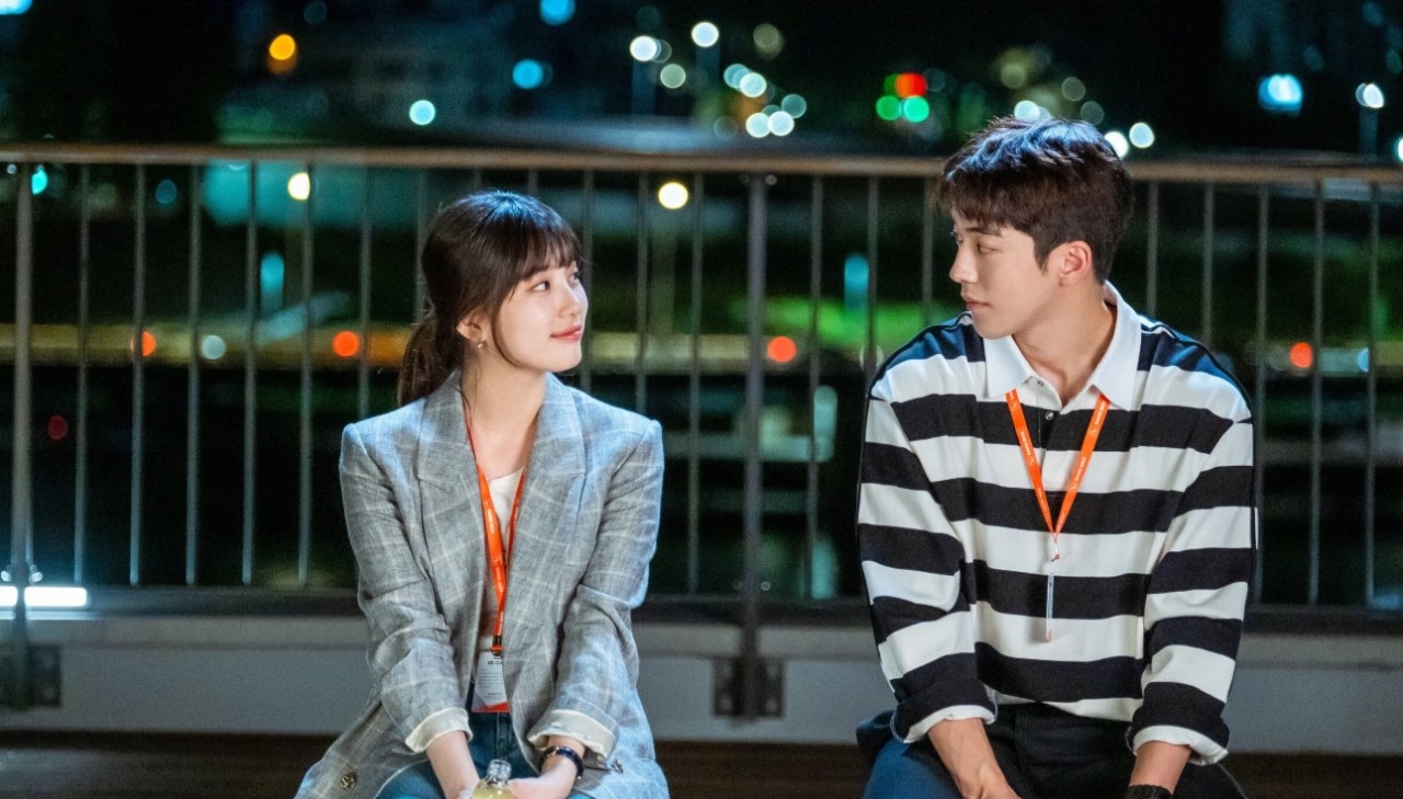 A sweet moment from *Start-Up*, featuring Seo Dal-mi and Nam Do-san sitting together on a balcony at night, gazing at each other with warm expressions, as city lights create a romantic atmosphere in the background.
