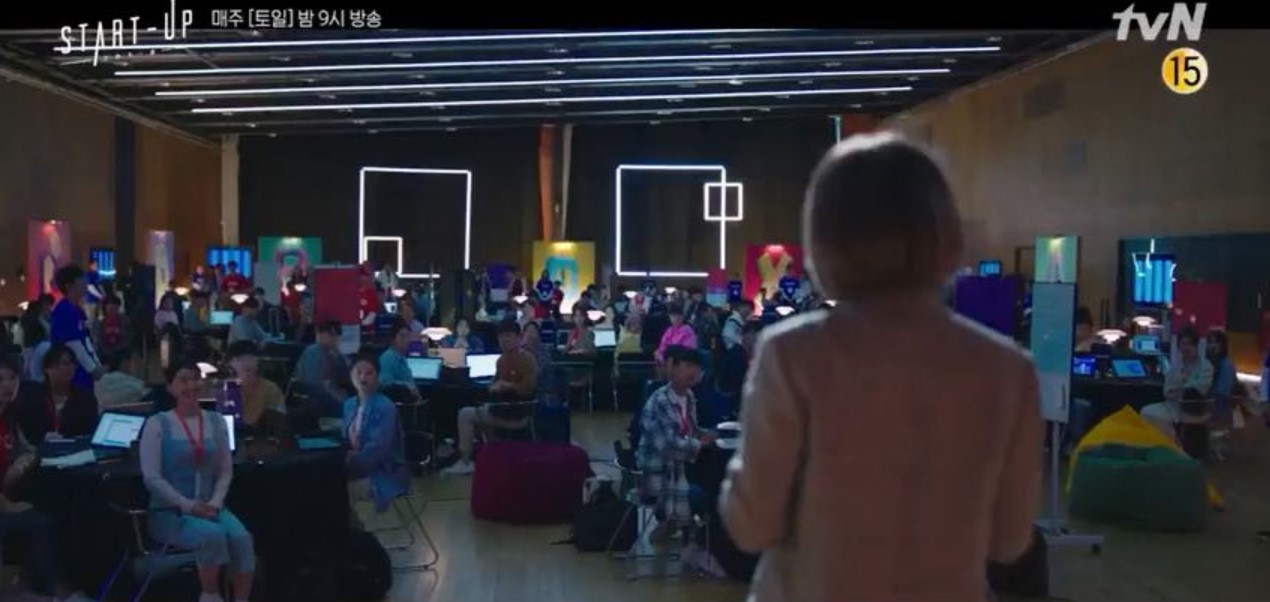 A vibrant scene from *Start-Up*, showing a large group of people working in a high-tech innovation space, with colorful decorations and multiple laptops. The focus is on a speaker standing at the front, addressing the crowd, setting the stage for a dynamic start-up environment.