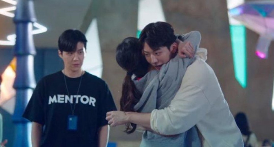 A dramatic scene from *Start-Up*, where Nam Do-san is hugging Seo Dal-mi while Han Ji-pyeong watches them from a distance, wearing a "MENTOR" shirt, highlighting the tension and complex relationships between the characters.
