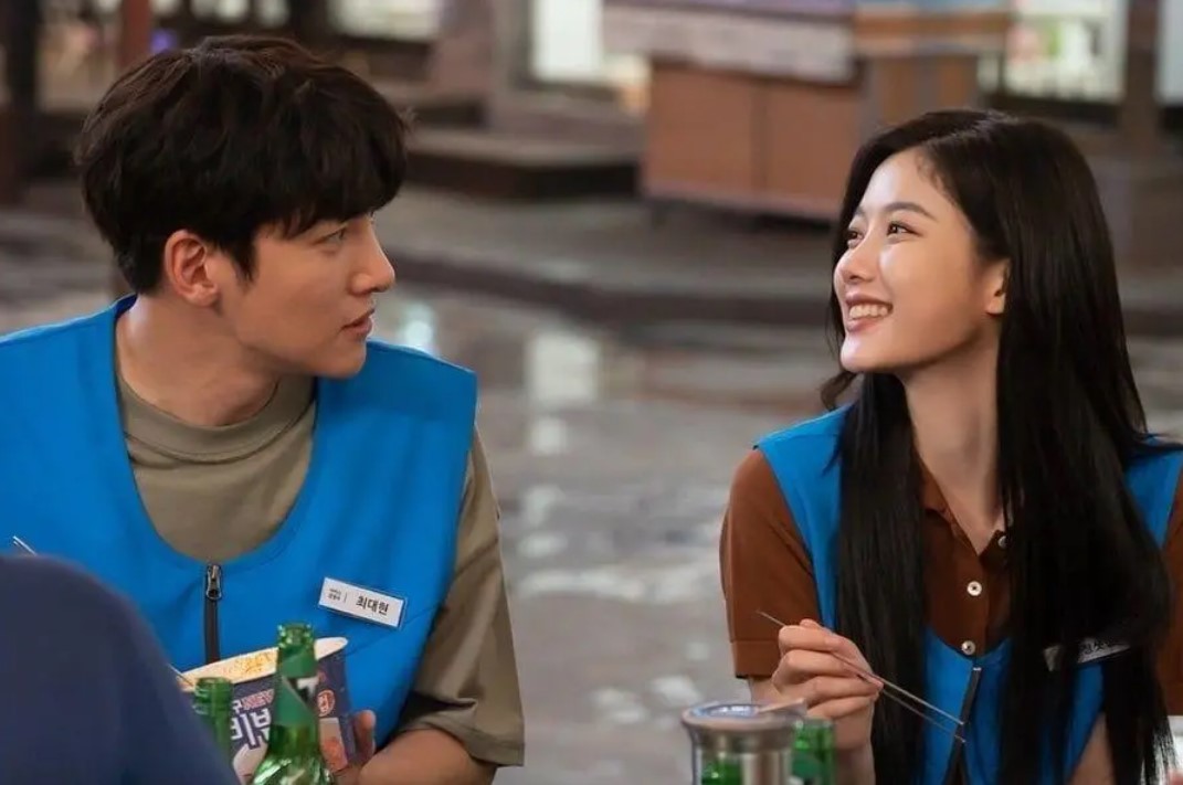 Ji Chang-Wook and Kim You-Jung sharing a heartwarming moment in 'Backstreet Rookie.' The two characters, Choi Dae-Hyun and Jung Saet-Byul, smile at each other while seated at a table in their convenience store uniforms, showcasing their growing bond in this romantic K-drama scene.