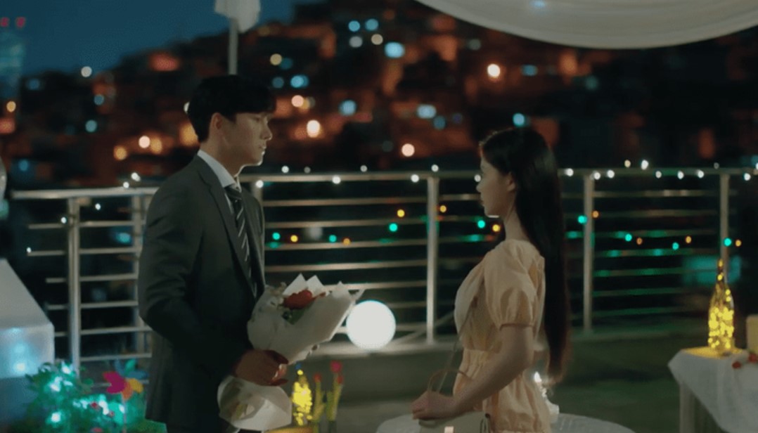 Ji Chang-Wook and Kim You-Jung in a romantic rooftop scene from 'Backstreet Rookie,' where Ji Chang-Wook's character, Choi Dae-Hyun, surprises Kim You-Jung's character, Jung Saet-Byul, with a bouquet of flowers. The beautiful nighttime setting, filled with soft lights and a cityscape background, adds to the heartfelt moment between the characters.