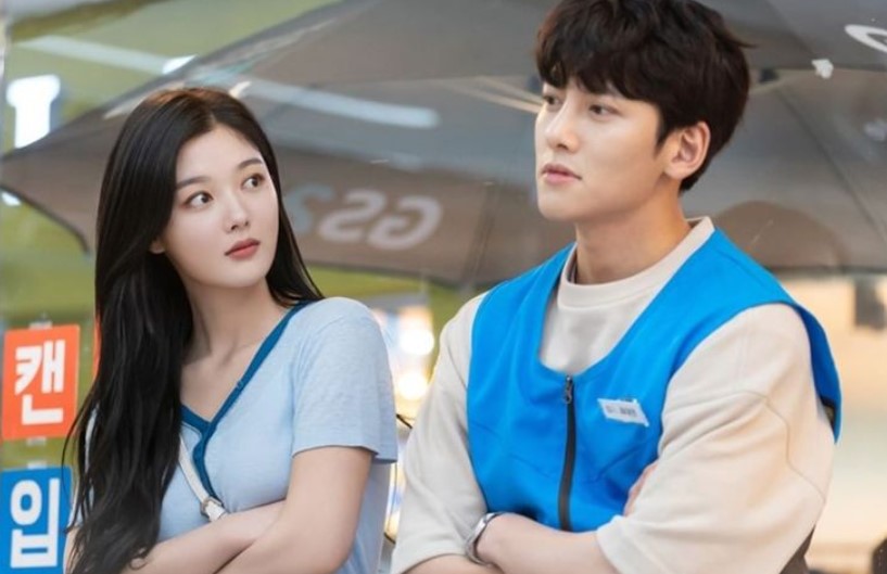 Ji Chang-Wook and Kim You-Jung in a playful moment from 'Backstreet Rookie,' as their characters, Choi Dae-Hyun and Jung Saet-Byul, exchange glances under an umbrella outside the convenience store. Both are in their work uniforms, with Saet-Byul looking curiously at Dae-Hyun, showcasing the lighthearted dynamic between the two leads.