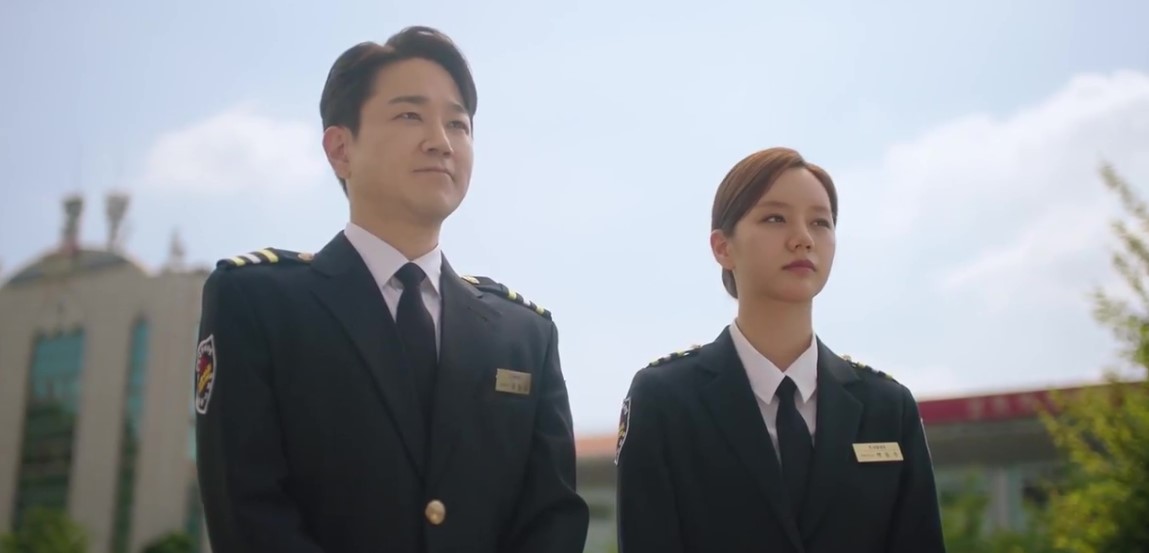 Two characters in uniform, standing side by side outside, under a clear sky, from a Korean drama.