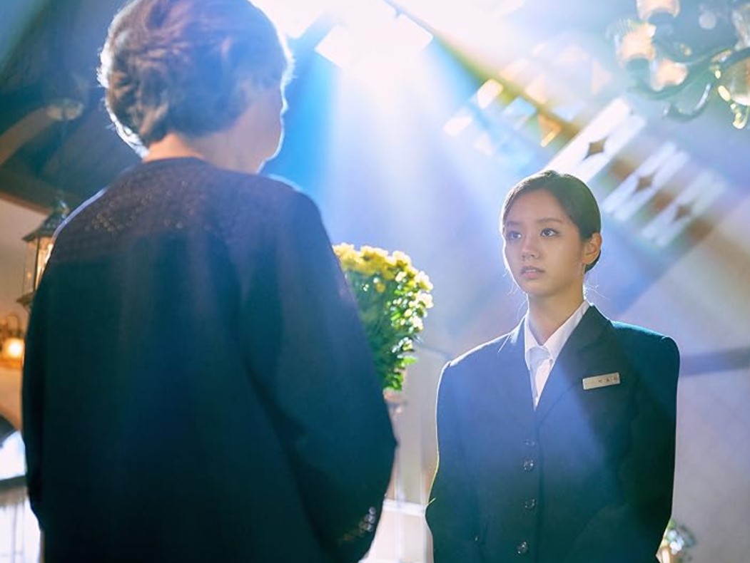 Young woman in a suit standing in a dramatic, light-filled scene with an elderly person holding flowers, from a Korean drama.