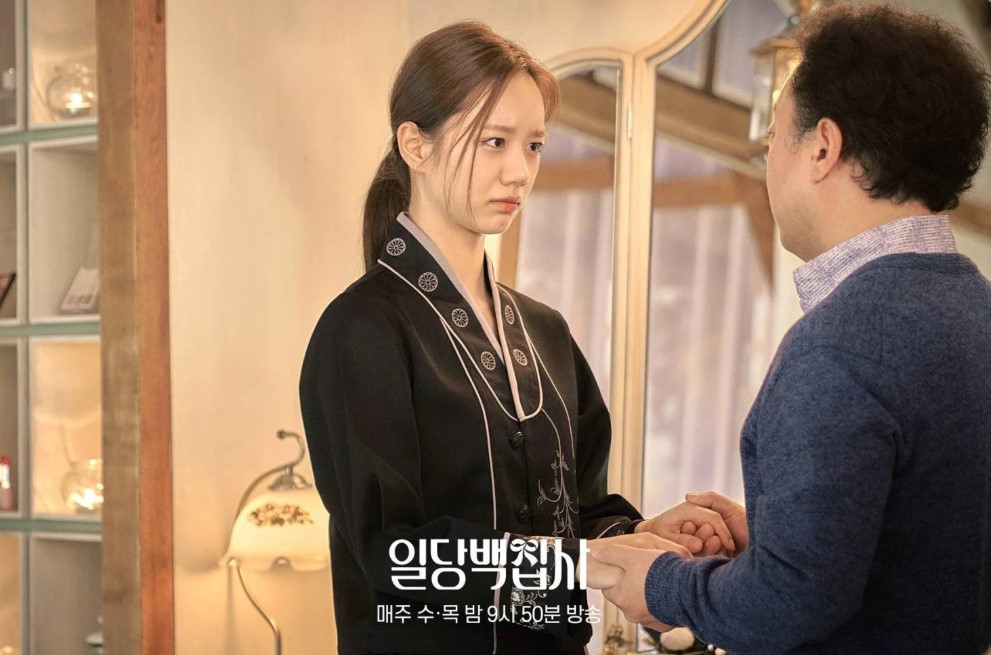 Emotional scene from Korean drama 'May i help you' featuring the female lead in traditional attire holding hands with an older man, conveying a moment of seriousness and concern.