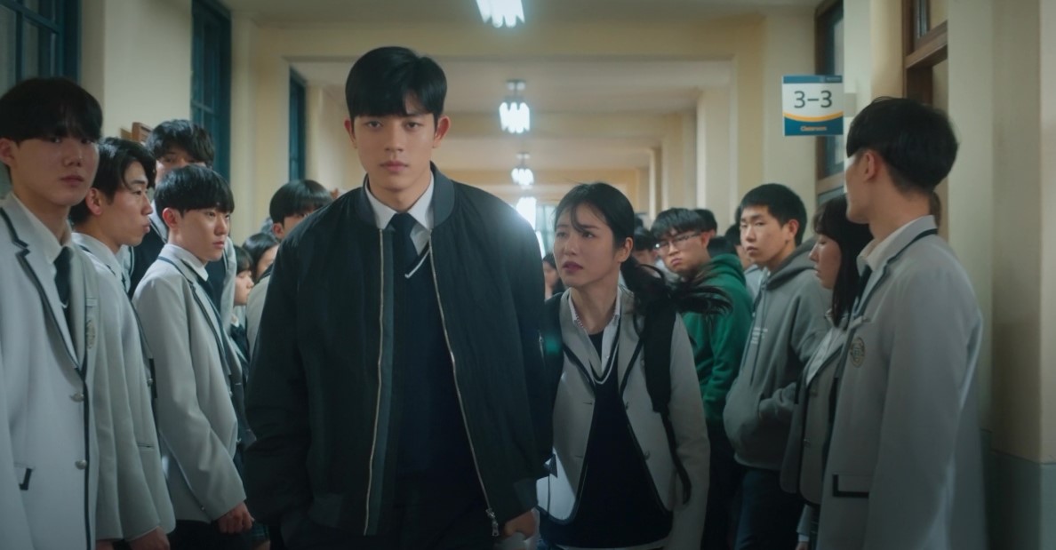 Two main characters from 'Revenge of Others' walking through a school corridor, surrounded by classmates in school uniforms, with the male character in a jacket and the female character following closely behind.
