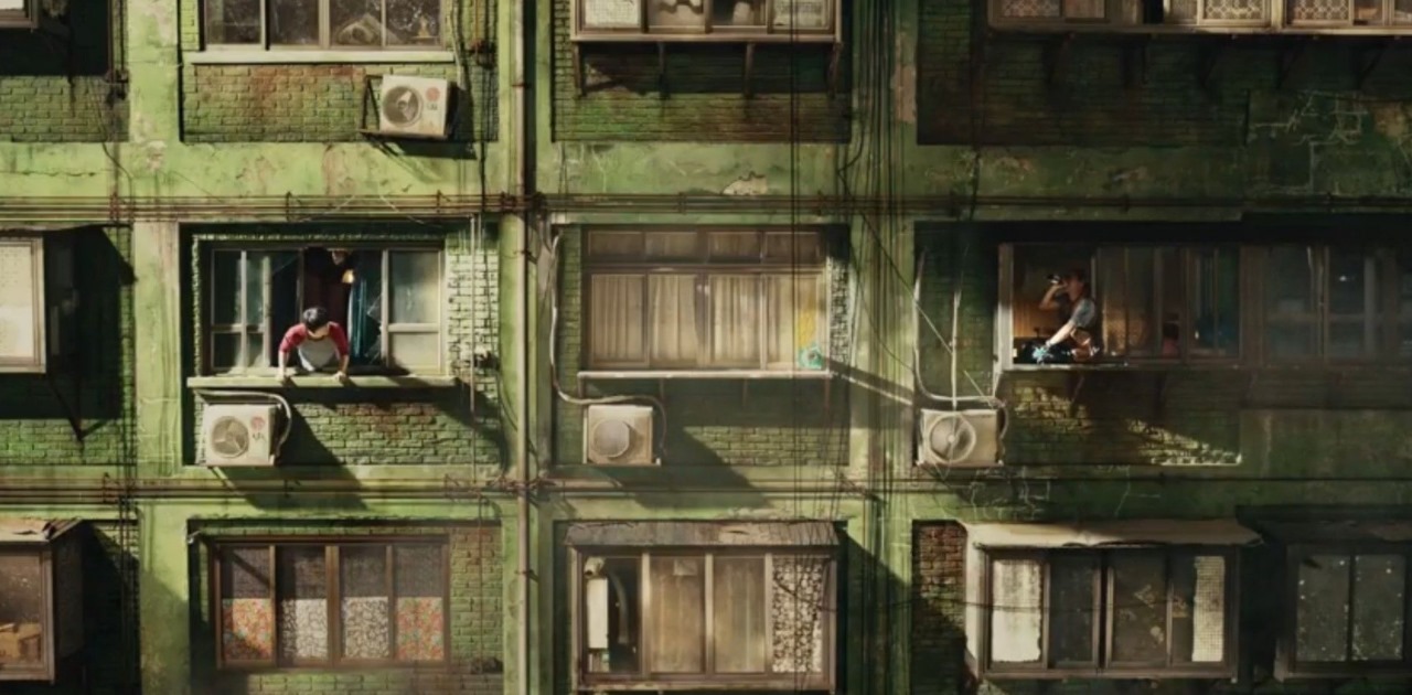 Wide shot of a green, run-down apartment building with characters sitting on the windowsills, one hanging from a window while the other sits casually, depicting a tense or dramatic scene from Sweet Home.
