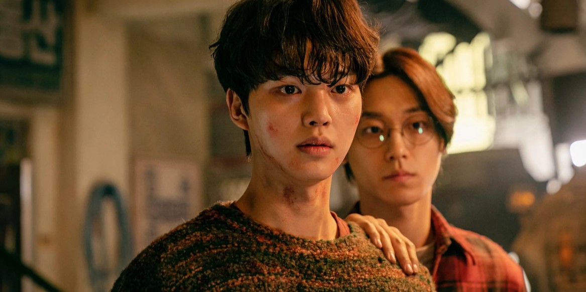 Intense moment from Sweet Home with a young man covered in dirt and bruises, standing in the foreground, while another character in glasses stands behind him, showing the aftermath of a dangerous situation.