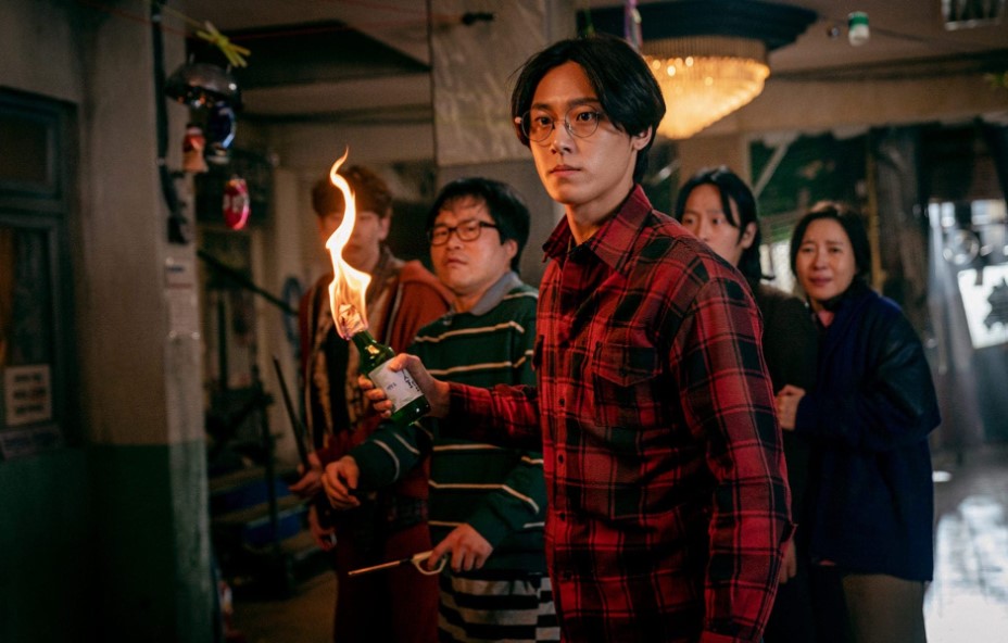 Character from Sweet Home holding a Molotov cocktail with a determined expression, surrounded by a group of people in the background, all preparing for a confrontation in a tense setting.