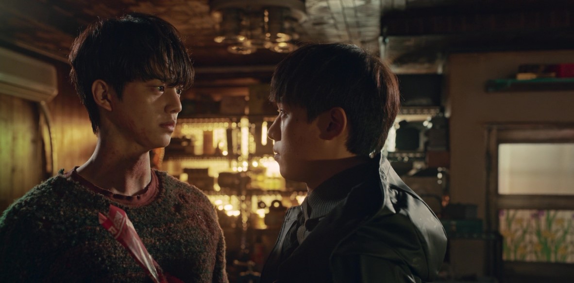Intense face-off between two characters from Sweet Home, with one holding a weapon and both staring each other down in a tense, dimly lit room.