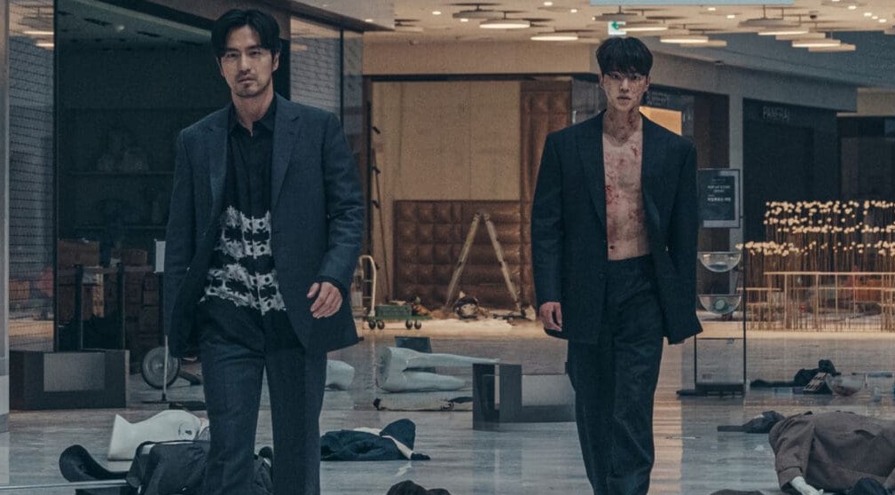 Two men walking confidently through a desolate mall, one in a dark suit with a bloodied chest and the other in a black shirt, surrounded by fallen bodies and chaos, creating a tense, post-apocalyptic scene.