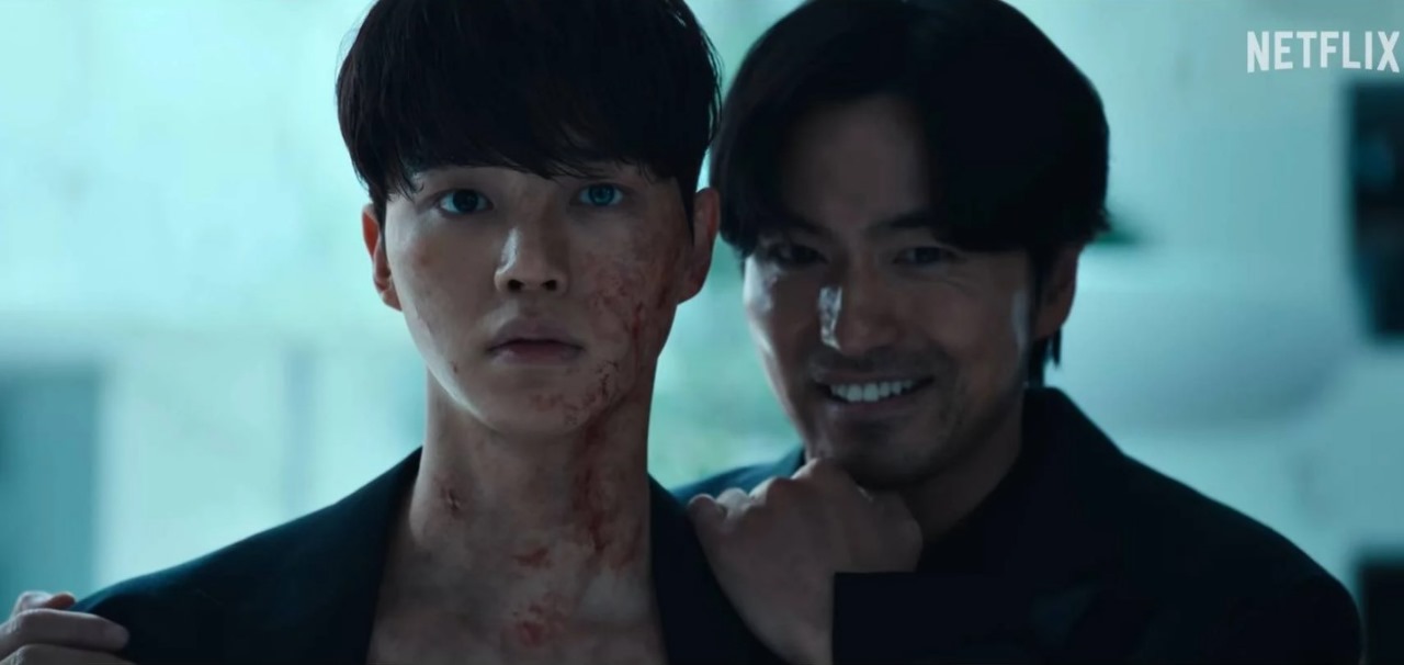 Intense close-up of a young man with scars on his neck, standing uneasily as a sinister figure smiles menacingly behind him, set in a tense, suspenseful atmosphere.