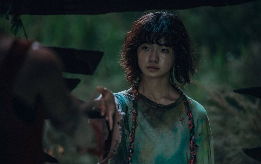 Young girl reaching out with a cautious expression, dressed in a tattered shirt, surrounded by a dark, forested environment with a dramatic, moody atmosphere.