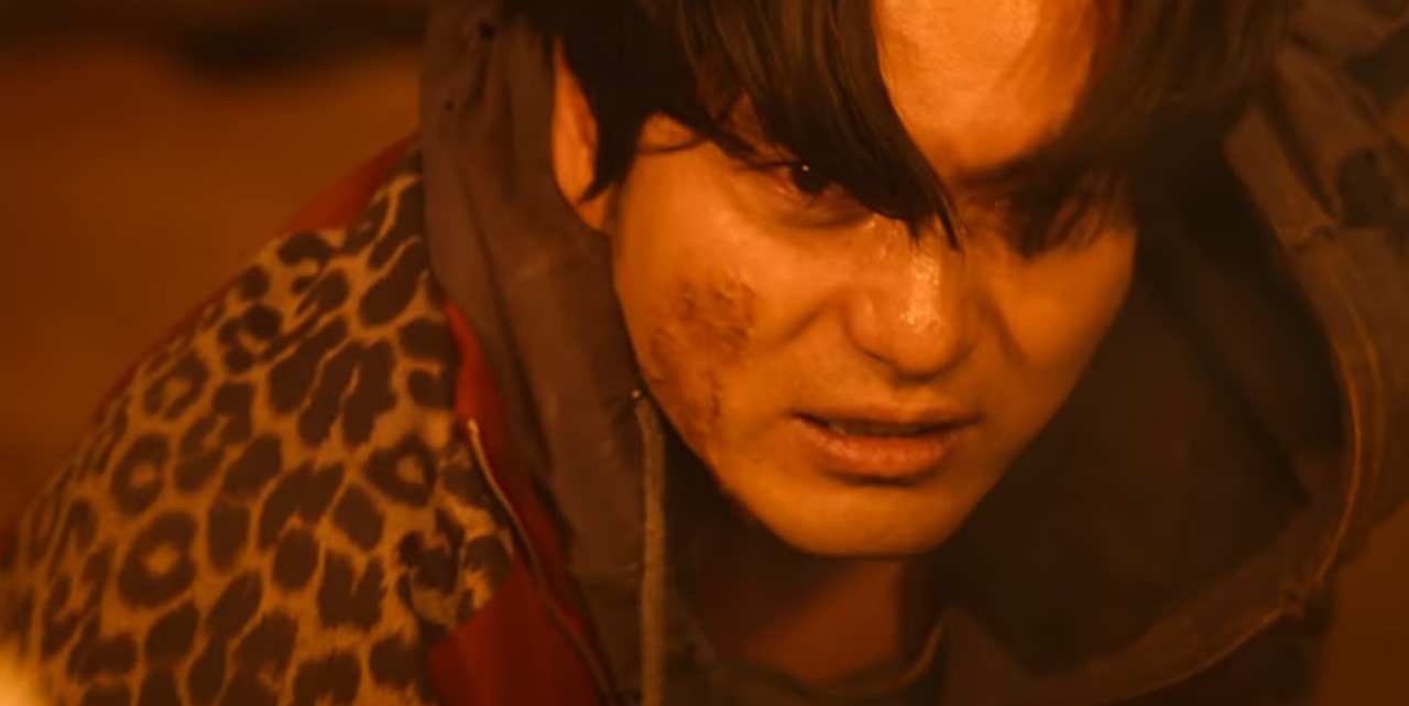 Close-up of a character from Sweet Home Season 2, showing a bruised and determined expression, with a leopard-print jacket, amidst a fiery orange backdrop.