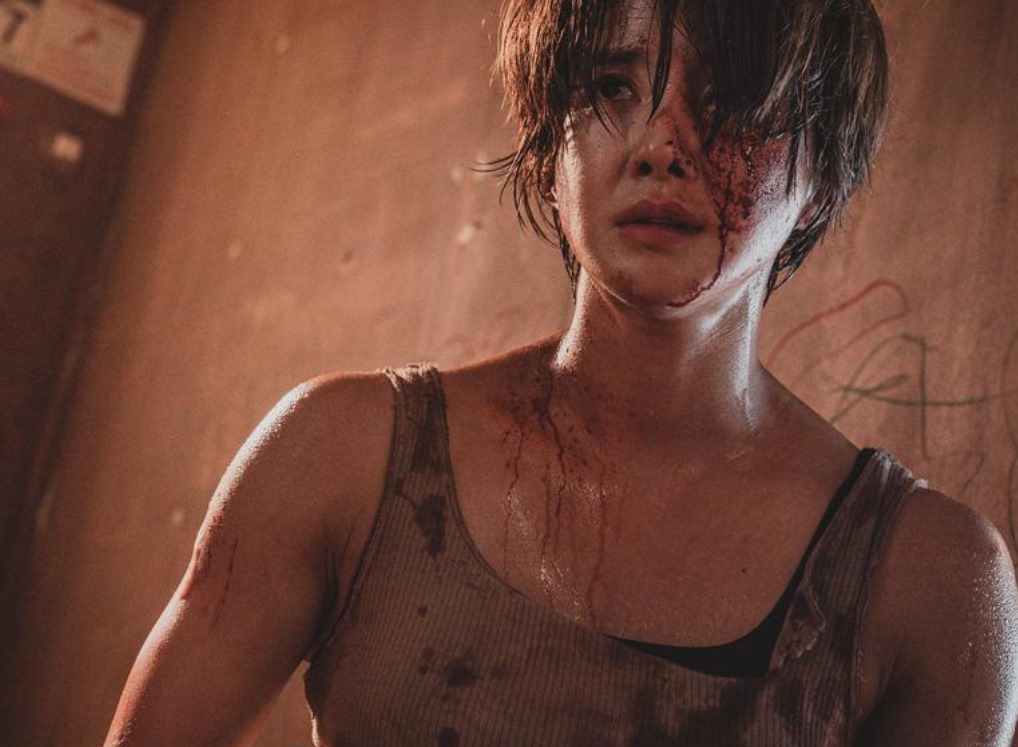 Close-up of a character from Sweet Home Season 2, covered in blood with a distressed expression, wearing a tank top, set against a grim, graffiti-covered wall.