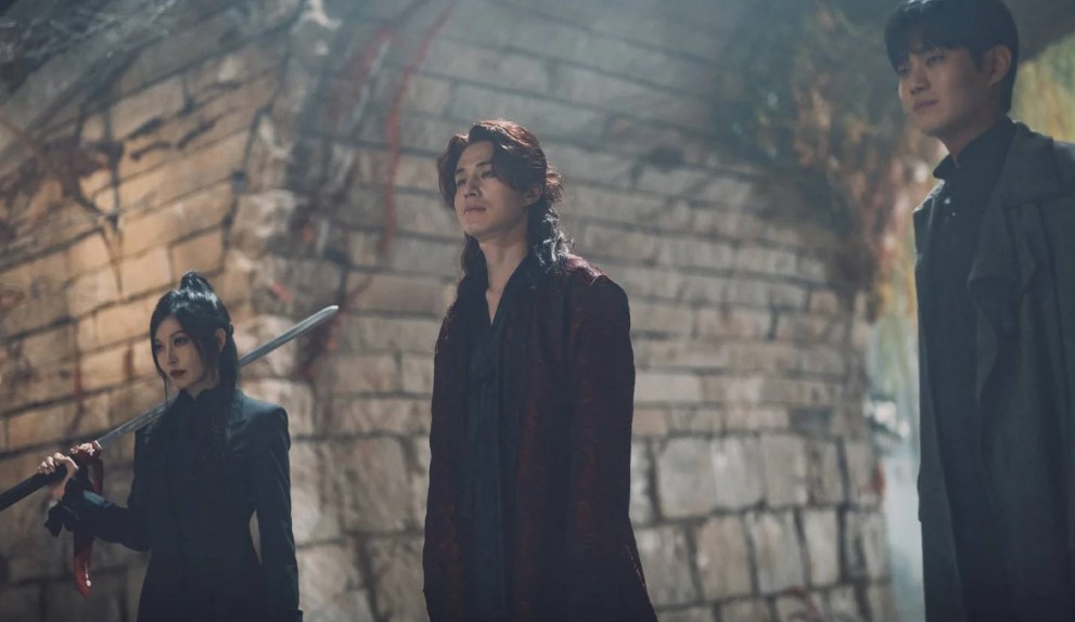 Scene from the South Korean drama 'Gumiho: 1985' showing three characters standing together in a dramatic pose inside a stone-walled structure, with the female character holding a sword and the two male characters in dark clothing.