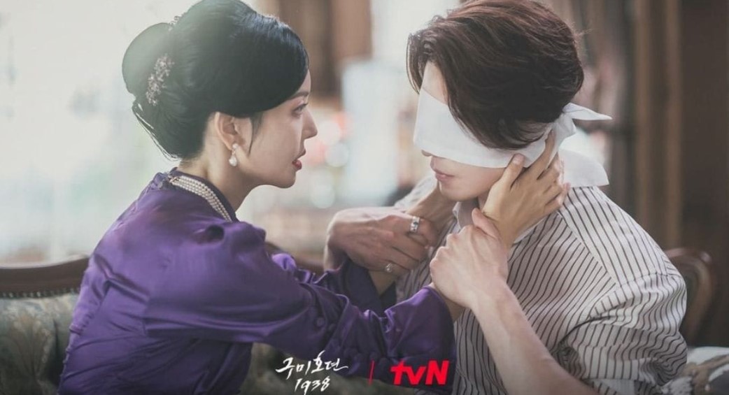 Romantic scene from the South Korean drama 'Gumiho: 1985' featuring a female character in a purple dress blindfolding a male character, with both of them in an intimate moment, set in an elegant indoor setting.