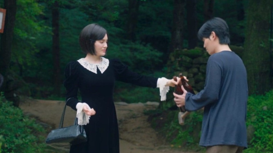Scene from the South Korean drama 'Gumiho: 1985' showing a female character in a vintage black dress handing a small doll to a male character in a forested area, with a calm and mysterious atmosphere.