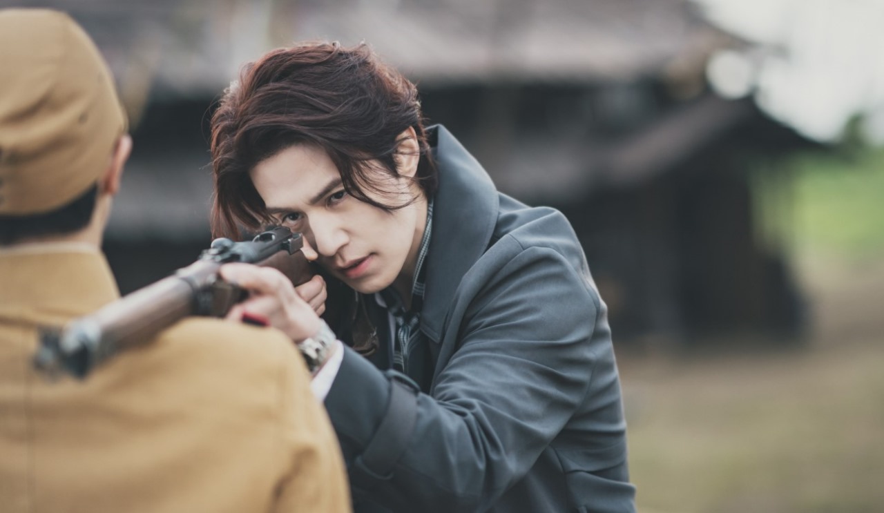 Scene from the South Korean drama 'Gumiho: 1985' featuring a male character aiming a rifle at another man in a tense standoff, set in a rural outdoor location with a vintage backdrop."