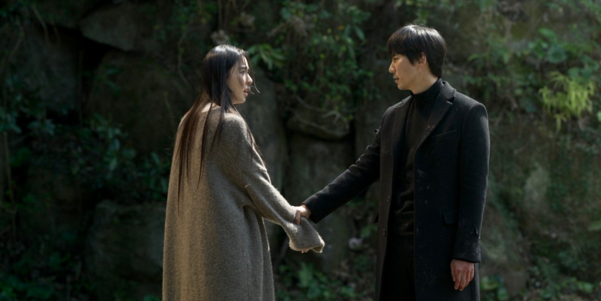Dramatic scene of a man and woman holding hands in a forest, both dressed in dark clothing, with emotional expressions and a tense atmosphere.