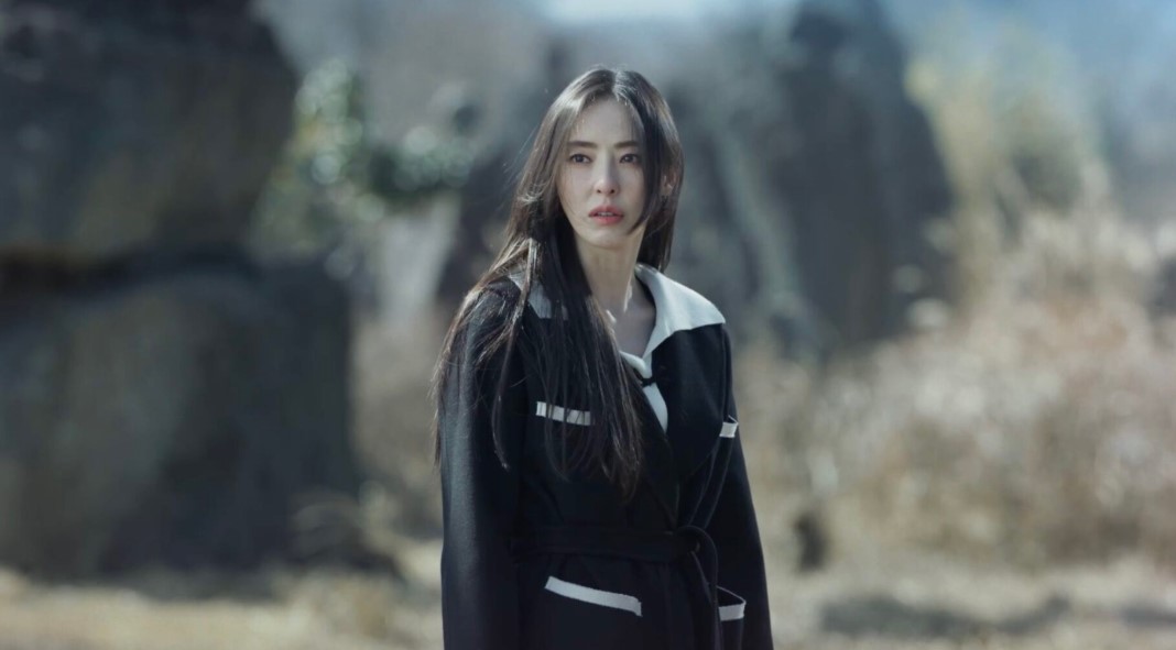 Woman in black coat standing in rocky landscape with intense expression