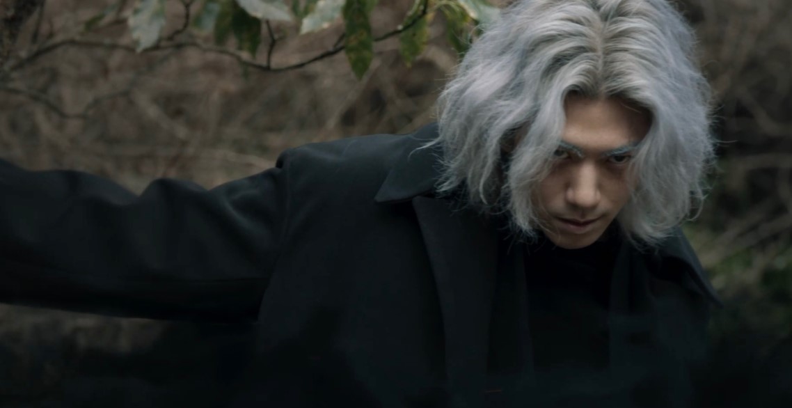 Character from 'Island' drama series with silver hair and intense gaze, dressed in a dark coat, standing in a forest setting, exuding a mysterious and menacing aura.