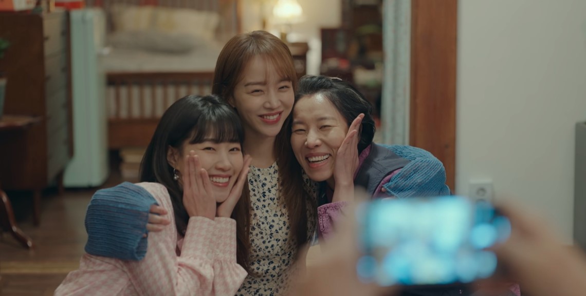 Heartwarming scene from 'This Life, Too, is Good,' featuring three female characters posing for a selfie, smiling and enjoying a joyful moment together in a cozy indoor setting.