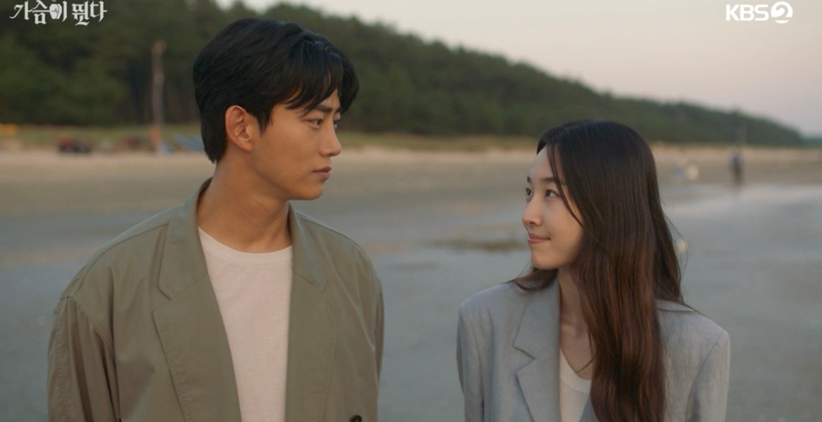 Emotional scene from 'Heartbeat' showing the main characters gazing at each other on a beach at sunset, with the man in a beige jacket and the woman in a light blue blazer, capturing a tender moment.
