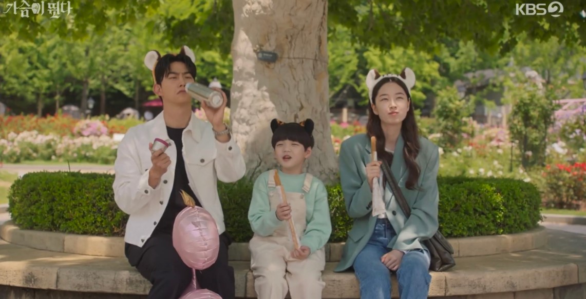 Adorable scene from Heartbeat showing the main characters and a child enjoying ice cream in a park, all wearing bear ears, creating a fun and heartwarming family moment.