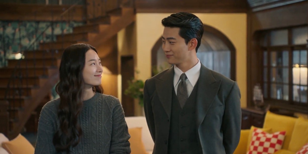 Romantic moment from the K-drama 'Heartbeat,' featuring the main characters smiling at each other in a cozy, elegant living room setting, with the woman in a sweater and the man in a sharp suit.