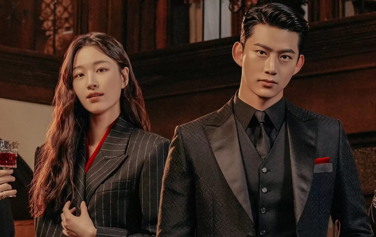 Main characters from the K-drama 'Heartbeat ' dressed in stylish suits, standing confidently in an elegant indoor setting. The woman holds a glass, while the man exudes a strong, charismatic presence.