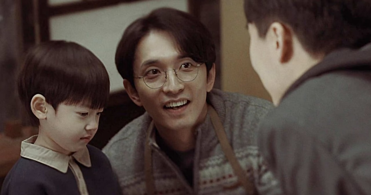 Scene from 'Miraculous Brothers' showing a warm moment between a smiling adult character wearing glasses and a young child. The child looks thoughtfully at another character, capturing a heartwarming family interaction.