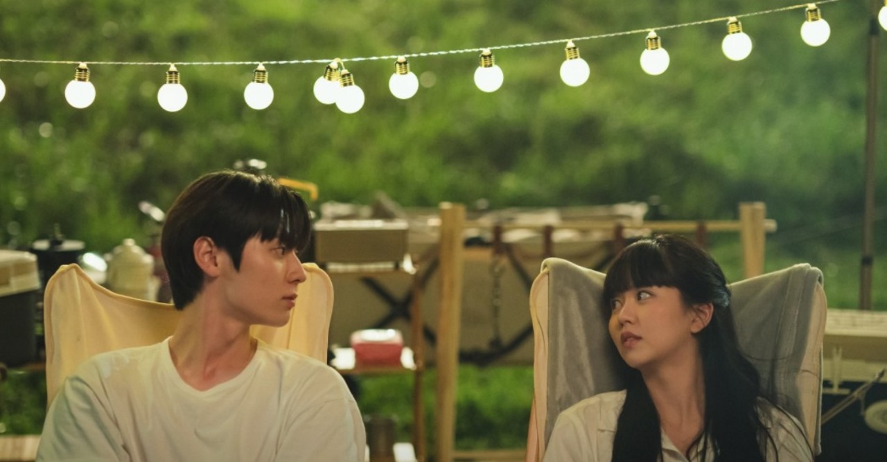 Scene from 'My Lovely Liar' showing the two main characters sitting across from each other under a string of lights, sharing a quiet and thoughtful moment in a peaceful outdoor setting.