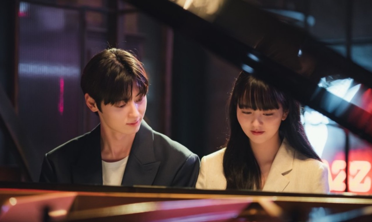 Scene from 'My Lovely Liar' showing two characters sitting at a piano, sharing a tender moment as they look at each other. The intimate setting captures the emotional connection between them.