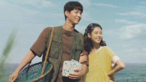 Two smiling characters in a scenic outdoor setting, holding a gift and fishing gear, featured in the Best K-Drama Reviews 2025. The scene captures a joyful moment between the characters in a romantic K-drama.