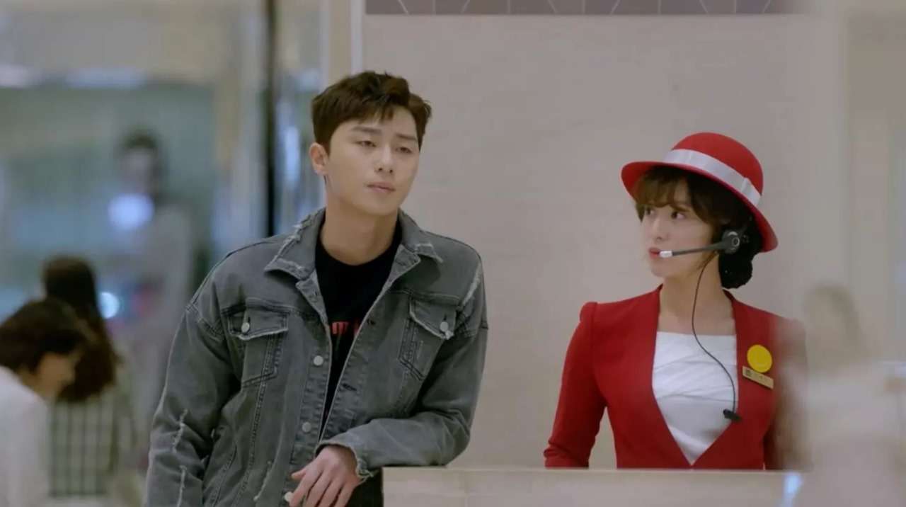 Park Seo-joon and Kim Ji-won in a playful scene from Fight for My Way, with Park Seo-joon dressed in a denim jacket and Kim Ji-won wearing a red uniform and headset as a mall worker. The moment captures their humorous and light-hearted interaction as their characters navigate romantic tension.